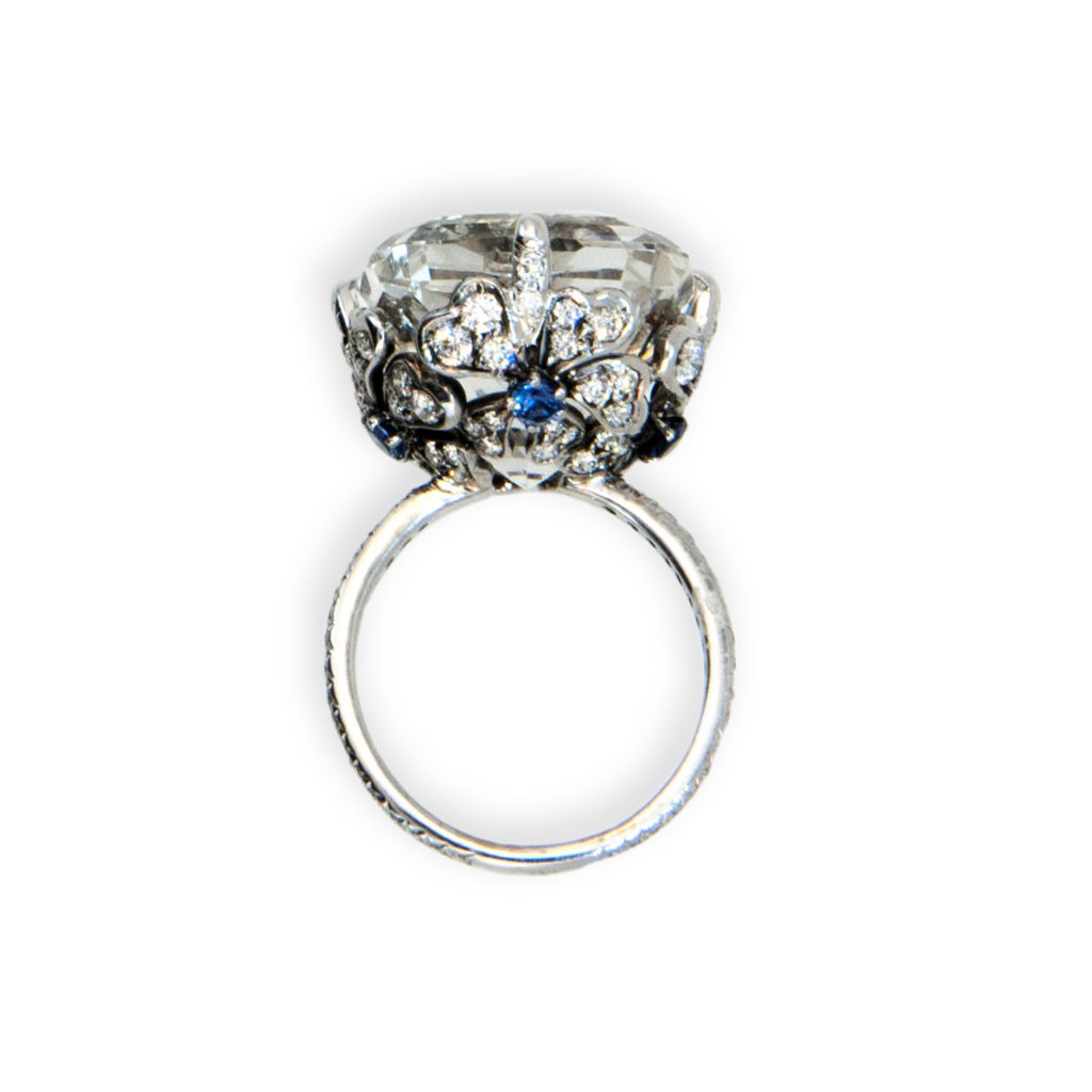 18 karat white gold ring set with one faceted pillow cut Topaz very light grey color 27.99 carats. Flower motif on sides of basket set with (4) round Blue Sapphires .32 carat total weight. Basket prongs and 1.6 mm shank are set with 104 round Diamonds 1.07 carats total weight. Ring size is 6 1/4.