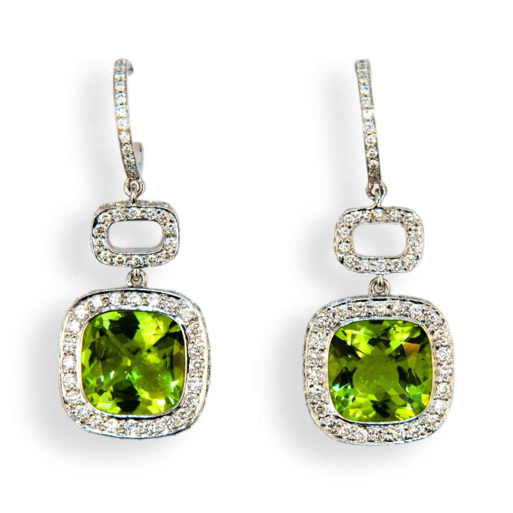 18 karat white gold earrings each set with one 10 x 10 mm cushion cut Peridot 3.36 carats and 3.66 carats. Set in each hoop are (18) diamonds .15 carat total weight. Surrounding Peridot and in connecting link are (85) diamonds 170 total approximately 1.02 carats total weight. For pierced ears.